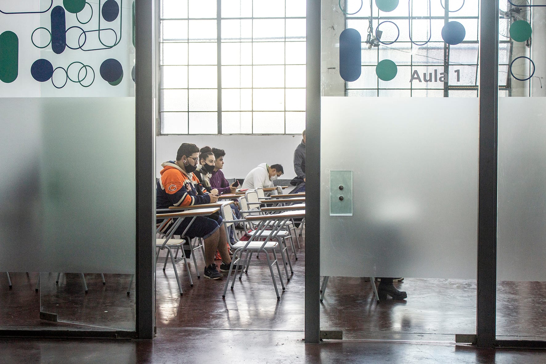 Estudiantes sentados en un aula, vistos a través de una puerta de vidrio.