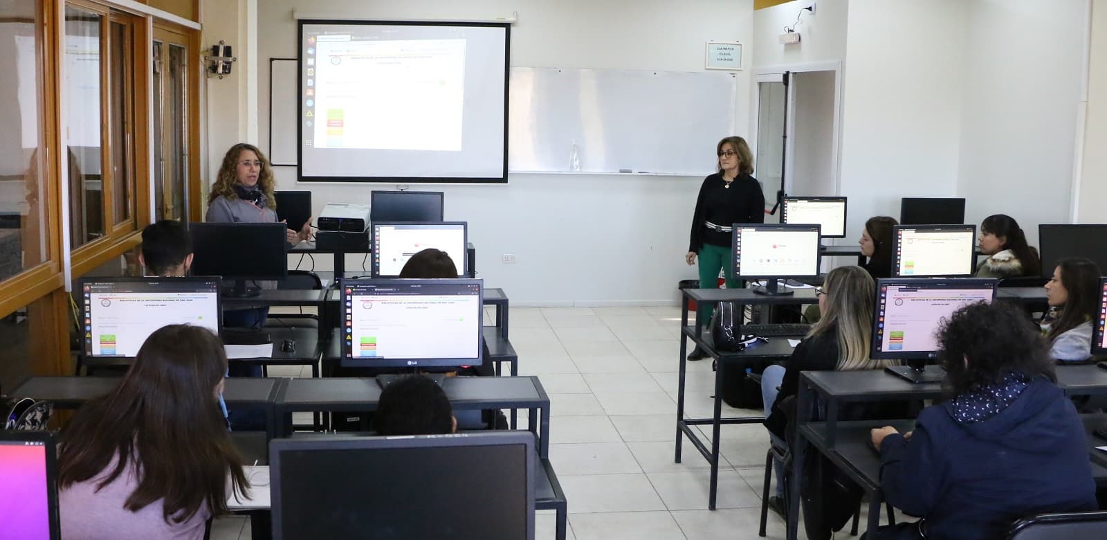 Aula informática con estudiantes frente a computadoras y dos docentes al frente