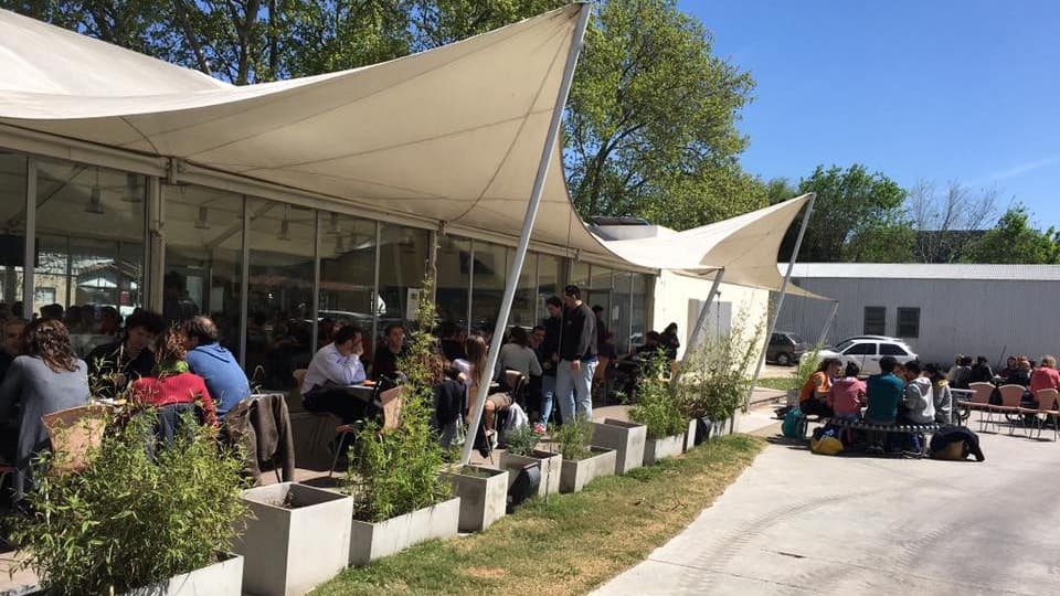 Grupos de estudiantes sentados en mesas y bancos al aire libre junto a un edificio de cubierta blanca.