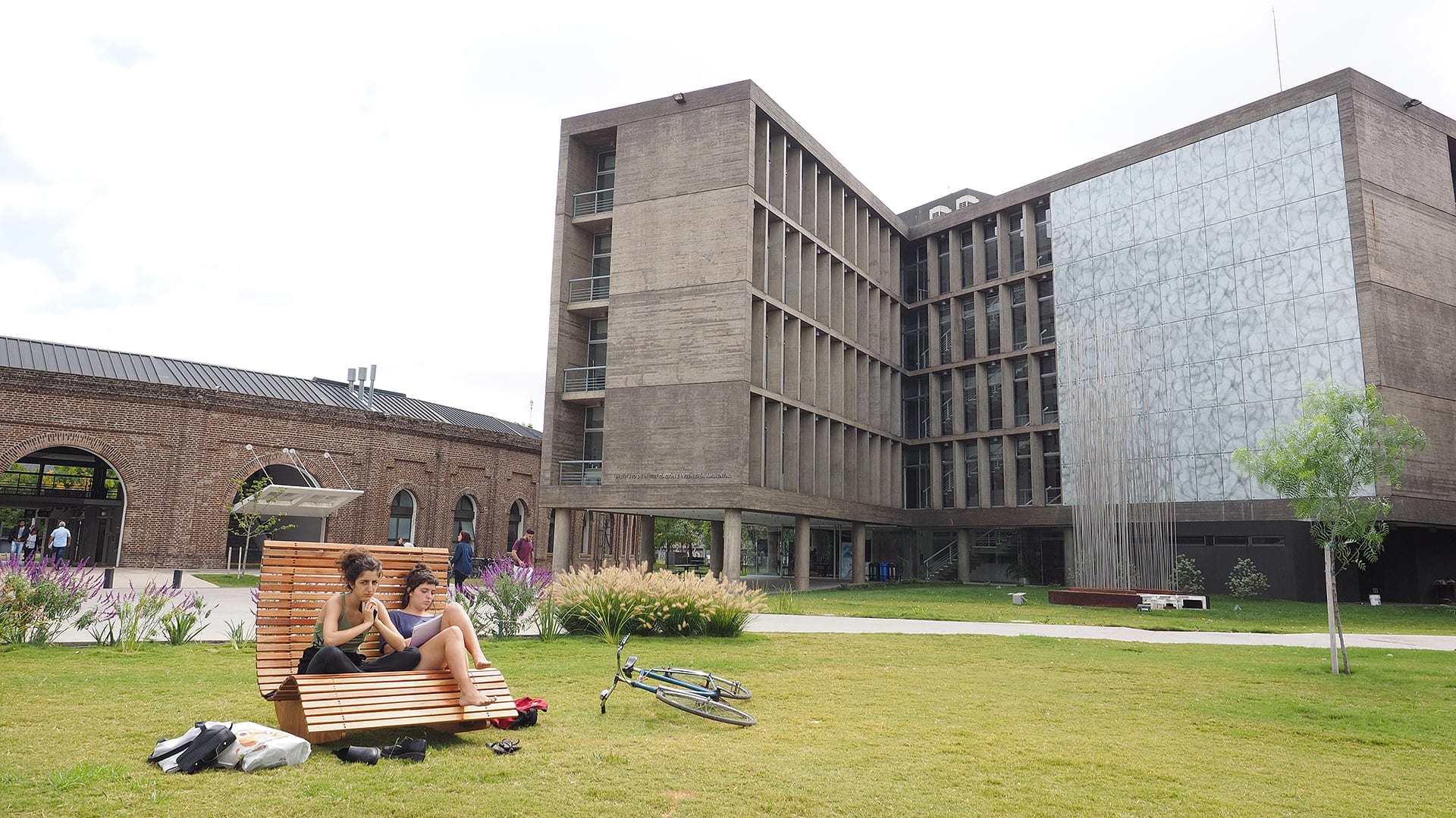 Edificio de varios niveles junto a un patio con césped, donde estudiantes están sentados al aire libre.