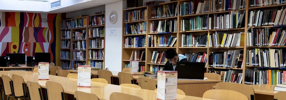 Sala de biblioteca con mesas de estudio, estanterías llenas de libros y una persona trabajando.