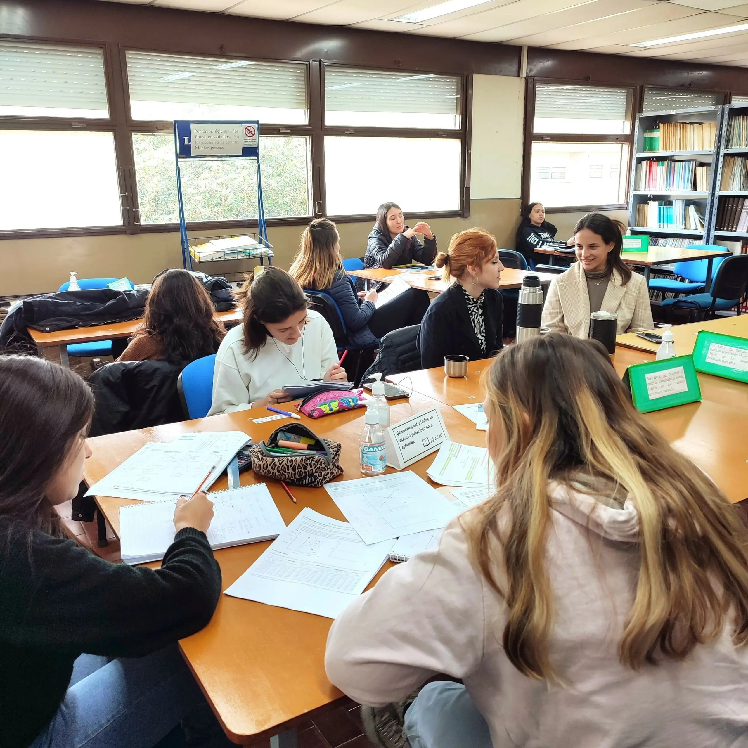 Estudiantes reunidas alrededor de mesas con apuntes en una sala de estudio con estanterías
