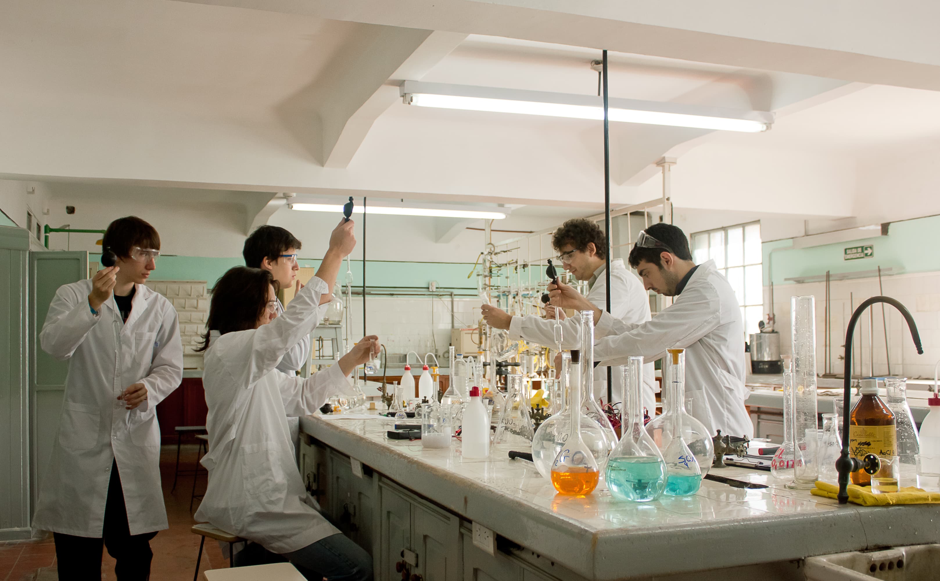 Estudiantes con guardapolvos realizan prácticas entre mesadas, matraces y equipos de laboratorio.