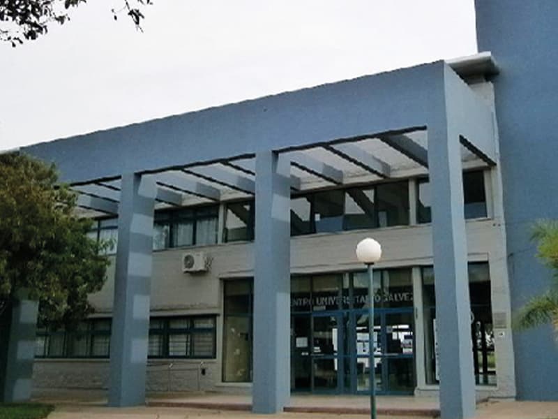 Fachada de un edificio universitario de dos plantas con acceso cubierto por columnas azules.