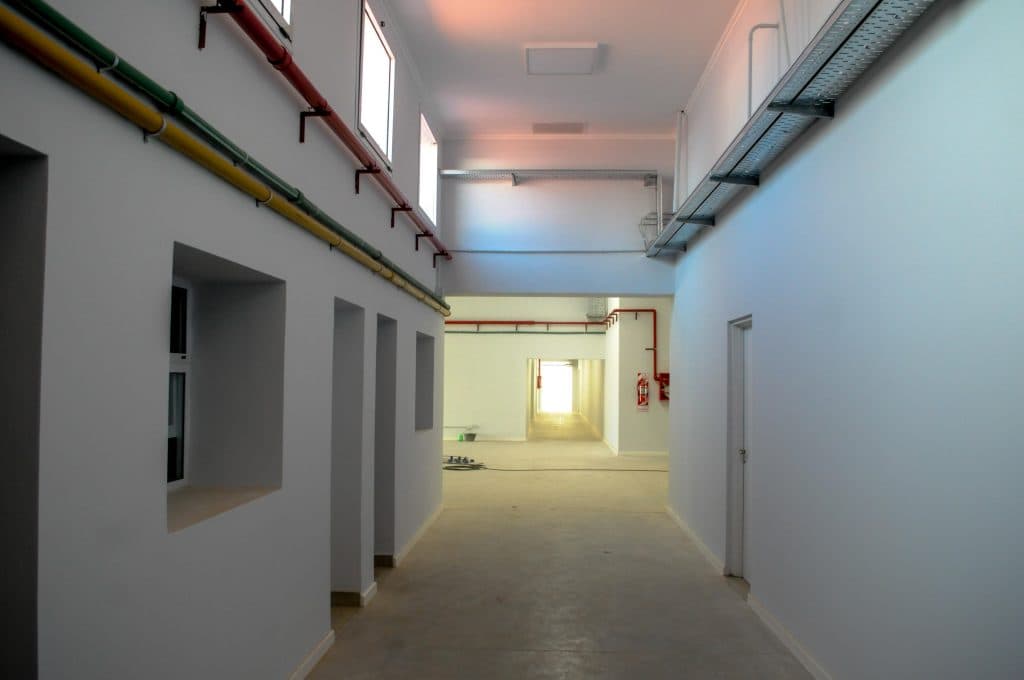 Pasillo interior amplio con puertas, ventanas altas e instalaciones a la vista.