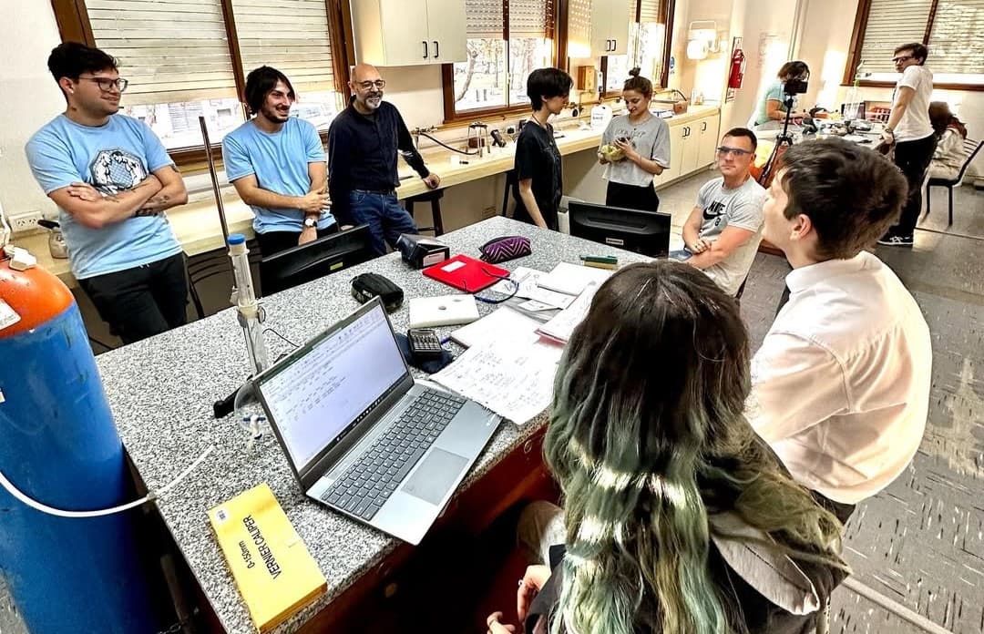 Estudiantes y docentes trabajan alrededor de una mesa con computadoras e instrumentos en un laboratorio de física.