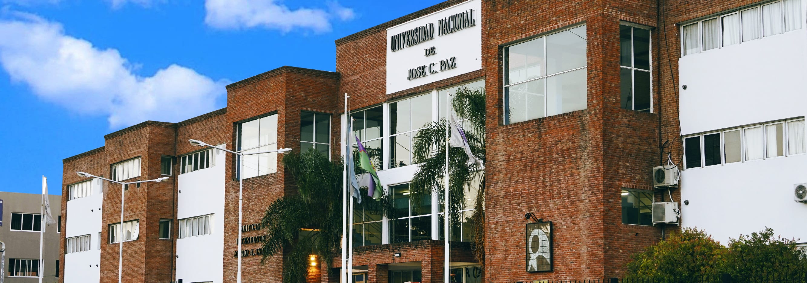 Fachada de ladrillo del edificio de la Universidad Nacional José C. Paz, con acceso central y banderas.